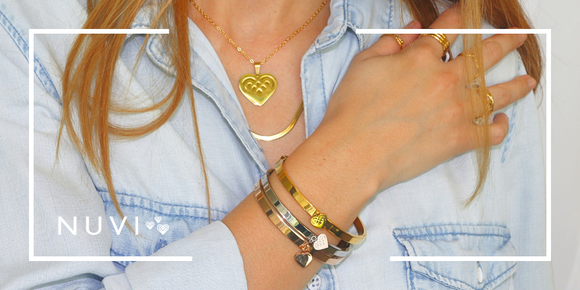 Mujer con camisa de mezclilla luciendo joyería dorada de NUVI ACCESORIOS: pulseras con dijes, anillos y collar con dije de corazón